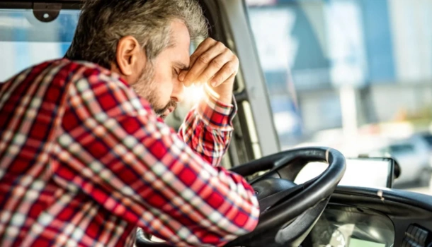 Frustrated truck driver in a red plaid shirt resting his head on the steering wheel inside the cab
