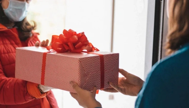 A person wearing a mask and red coat hands a festively wrapped gift box with a red bow to another person at a doorway