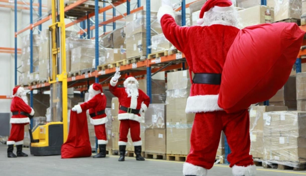 Multiple people dressed as Santa Claus organizing large red sacks in a warehouse filled with stacked pallets