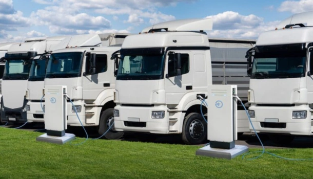 A row of white electric trucks is lined up and charging at EV stations on a grassy field