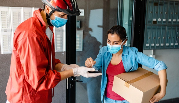 Delivery worker wearing a helmet and mask hands over a package to a masked customer
