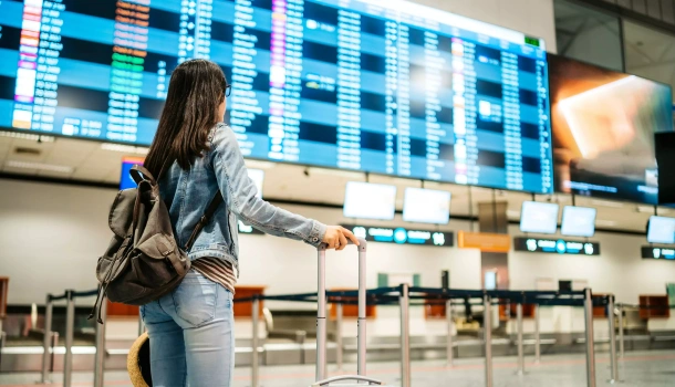 A woman with a backpack and suitcase stands in front of a large flight information board
