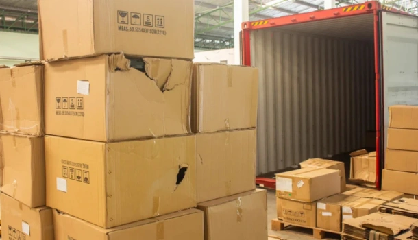 A stack of damaged cardboard boxes sits beside an open shipping container in a warehouse