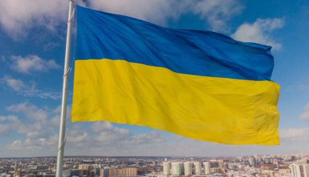 The Ukrainian flag waves prominently against a partly cloudy sky with a cityscape below