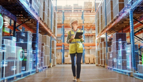 A warehouse worker in a safety vest and helmet walks through a digitally enhanced storage area with a tablet