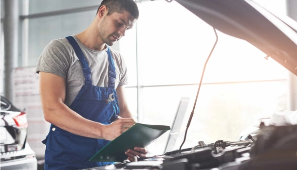 A male mechanic in blue overalls inspects the engine of a vehicle with the hood open