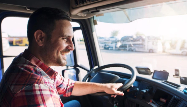 A smiling male truck driver in a plaid shirt sits behind the wheel of his truck