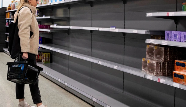 A woman holding a shopping basket stands in front of nearly empty supermarket shelves