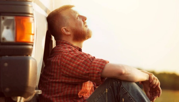 A tired HGV driver in a plaid shirt resting against his truck under the summer sun