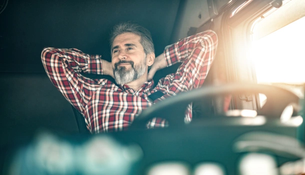 Smiling male truck driver in a red plaid shirt relaxing with hands behind his head inside a truck cabin