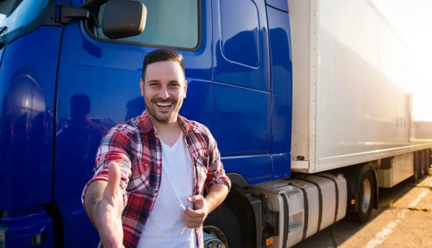 A smiling truck driver extends a handshake while standing in front of a blue semi-truck