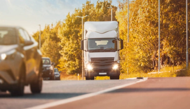 A white delivery truck drives on a sunlit road surrounded by cars