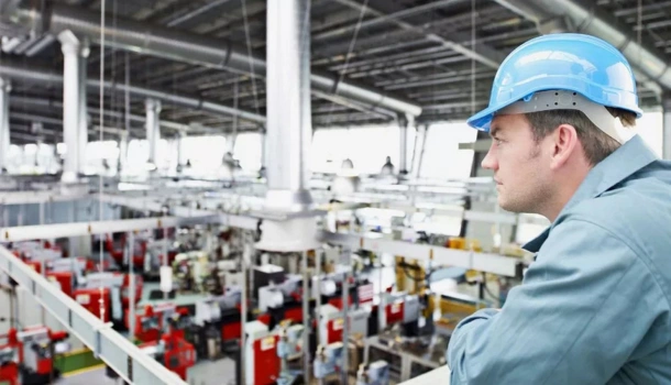 A man wearing a blue hard hat and work uniform overlooks a large, modern manufacturing facility