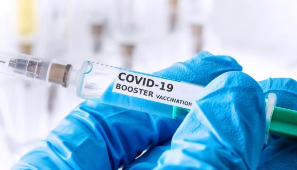 A gloved hand holds a syringe labeled "COVID-19 Booster Vaccination"