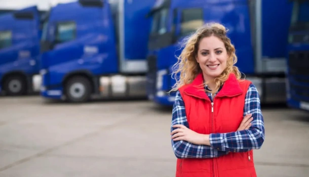 Woman in red vest and plaid shirt smiling confidently in front of a row of parked blue transport trucks